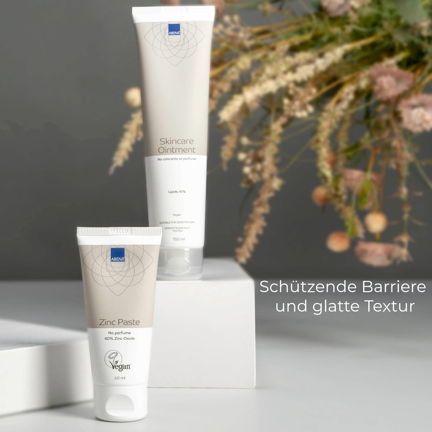 Zwei Pflegeprodukte stehen auf einem weißen Podest: eine Tube mit -Skincare Ointment- (150 ml) und eine mit -Zinc Paste- (60 ml). Im Hintergrund sind Blumen sichtbar. Text: -Schützende Barriere und glatte Textur-.