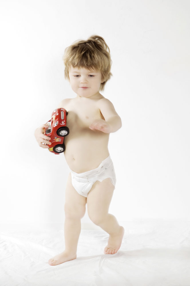 Ein kleines Kind mit kurzen Haaren trägt eine Windel und hält ein rotes Spielzeugauto in der Hand. Es steht auf einem weißen Untergrund und schaut freundlich dabei.