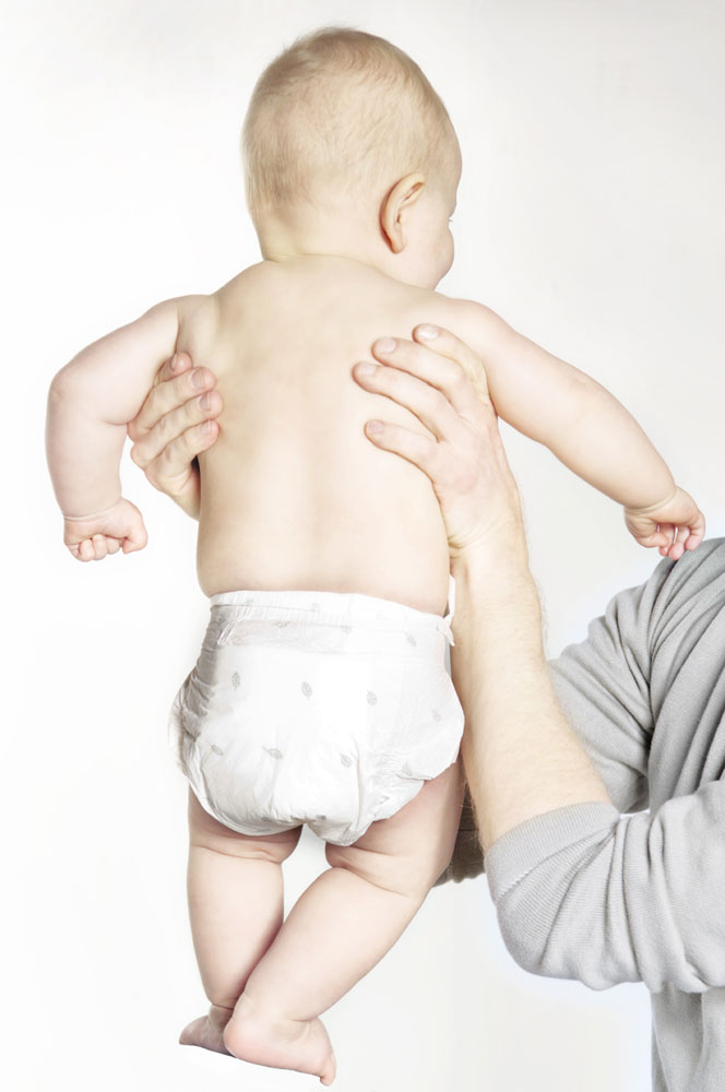 Ein Baby wird durch einen Erwachsenen hochgehoben, während es eine hellgraue Windel mit Punkten trägt. Der Hintergrund ist hell und neutral. Die Arme des Erwachsenen sind sichtbar.