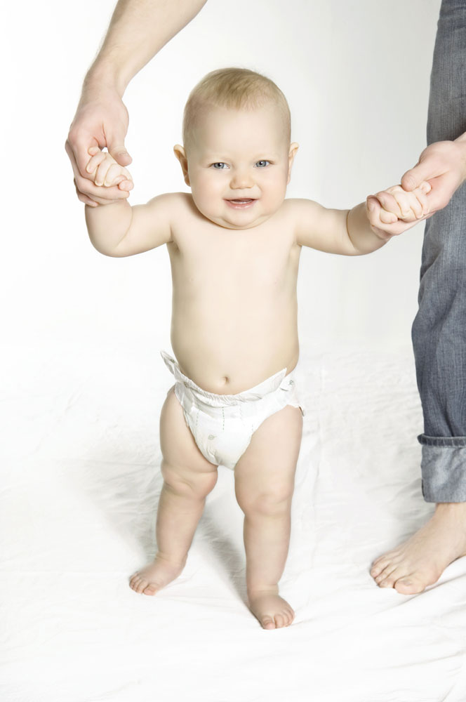 Ein Baby steht aufrecht und hält die Hände eines Erwachsenen. Es trägt eine weiße Windel und lächelt. Der Hintergrund ist hell und neutral, mit einfacher heller Unterlage.